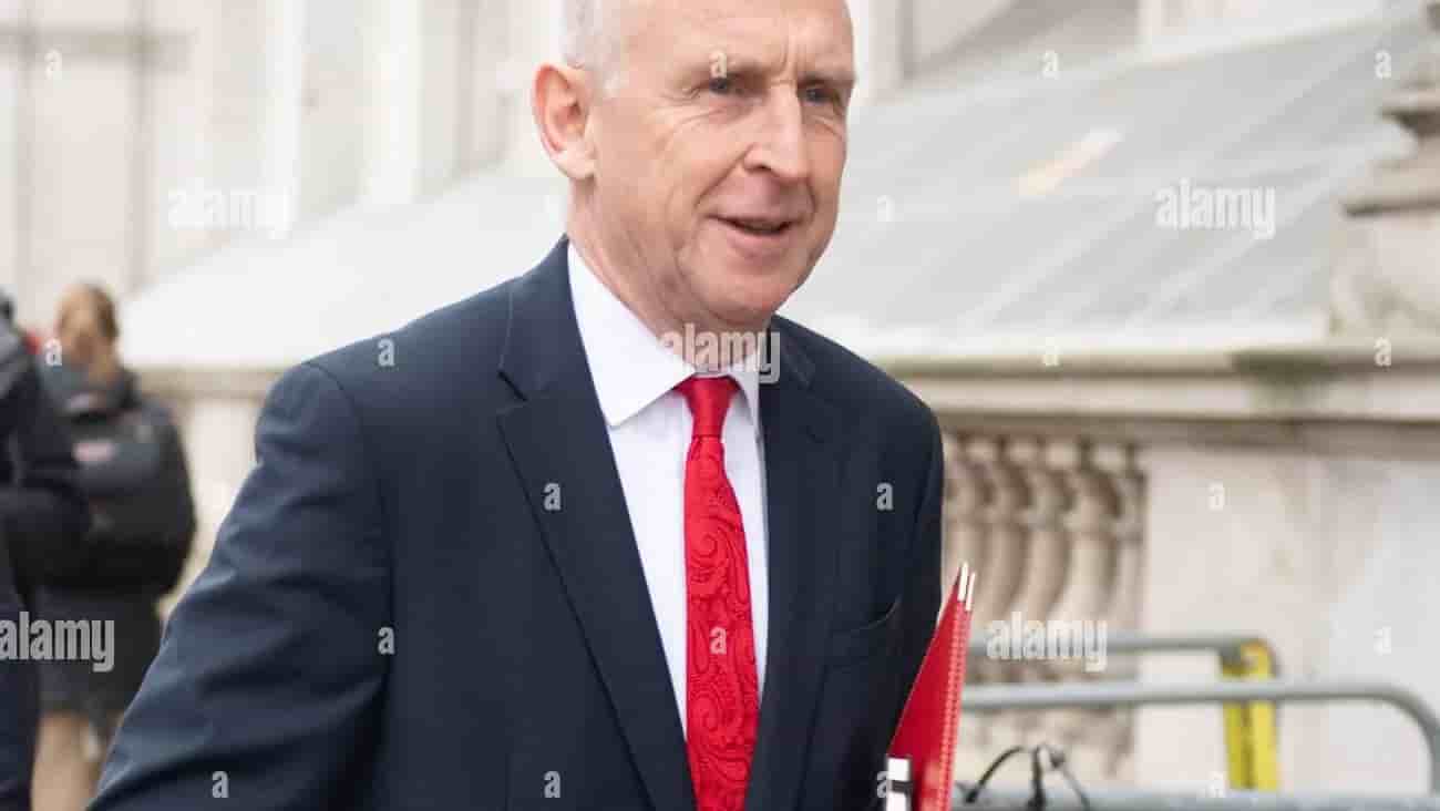 John Healey is smiling while wearing a dark suit and a red tie, holding red folders, as he walks past a building.